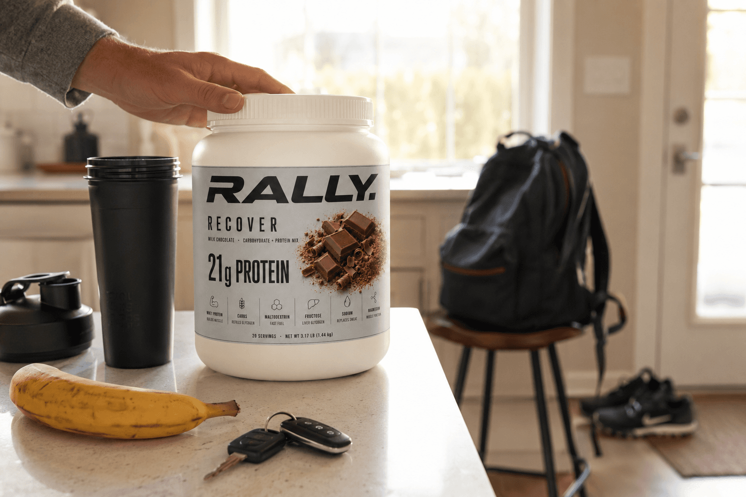 A hand resting on a tub of RALLY RECOVER on a kitchen counter, backpack and cleats behind